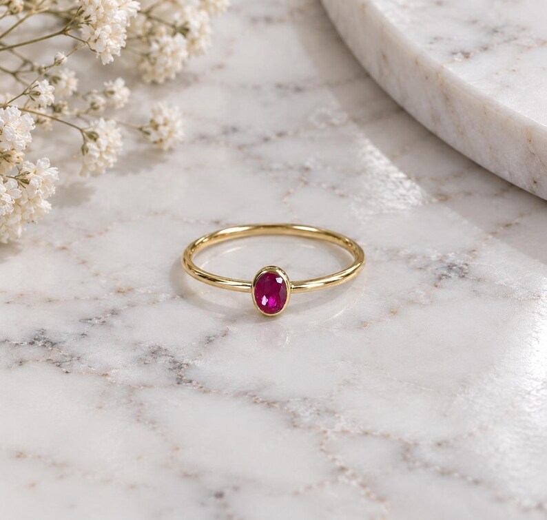 May include: A thin gold ring featuring a single, small, oval-shaped, vibrant pinkish-red gemstone set in a smooth bezel setting. The band is polished and slender, creating a delicate appearance for this piece of fine jewelry.