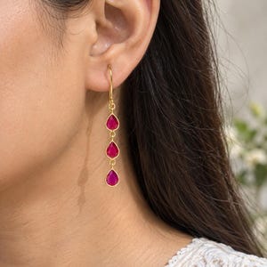 May include: A gold-toned drop earring featuring three vertically aligned, teardrop-shaped fuchsia pink stones. The earring hangs from a simple gold-toned hook wire, with each stone encased in a thin gold-toned metal bezel setting connected by small circular jump rings.
