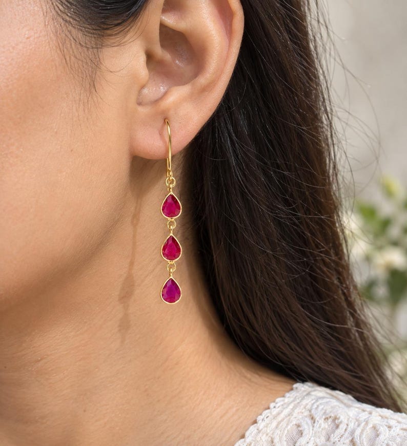May include: A gold-toned drop earring featuring three vertically aligned, teardrop-shaped fuchsia pink stones. The earring hangs from a simple gold-toned hook wire, with each stone encased in a thin gold-toned metal bezel setting connected by small circular jump rings.