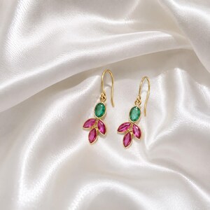 May include: A pair of gold drop earrings featuring a single oval green gemstone at the top, followed by three marquise-cut pink gemstones arranged in a cluster below, suspended from curved gold ear wires on a smooth white satin fabric background.