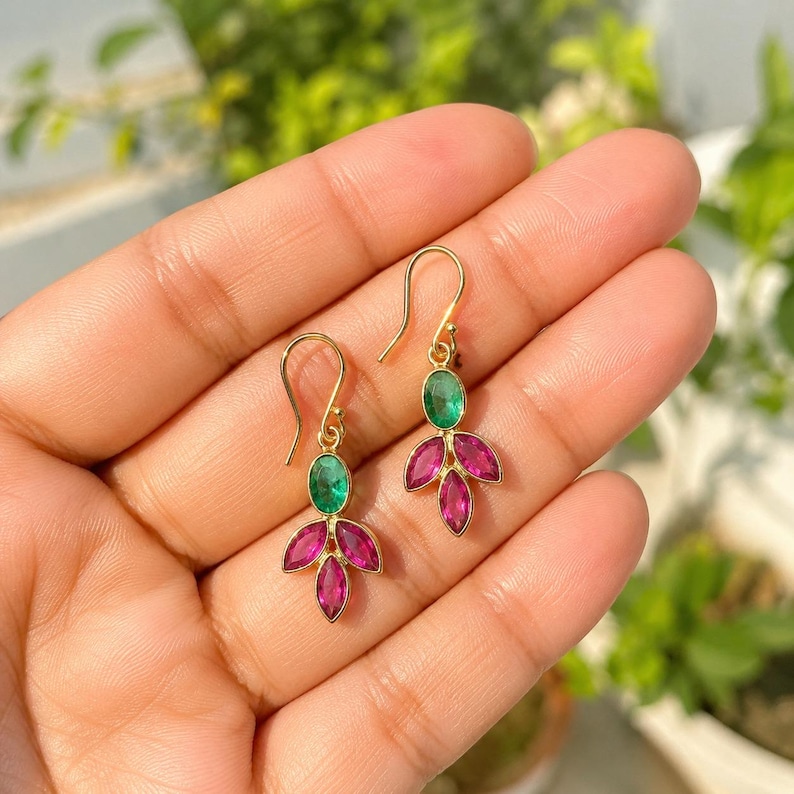 May include: A pair of gold-tone drop earrings featuring a single oval green gemstone at the top, accented by three marquise-cut pink gemstones arranged in a fan-like cluster below, suspended from simple curved gold-tone ear wires.