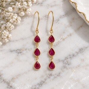 May include: A pair of gold drop earrings, each featuring three vertically aligned, teardrop-shaped, deep pinkish-red gemstones suspended from a curved gold ear wire. The stones are set in thin gold bezels and connected by small gold jump rings.