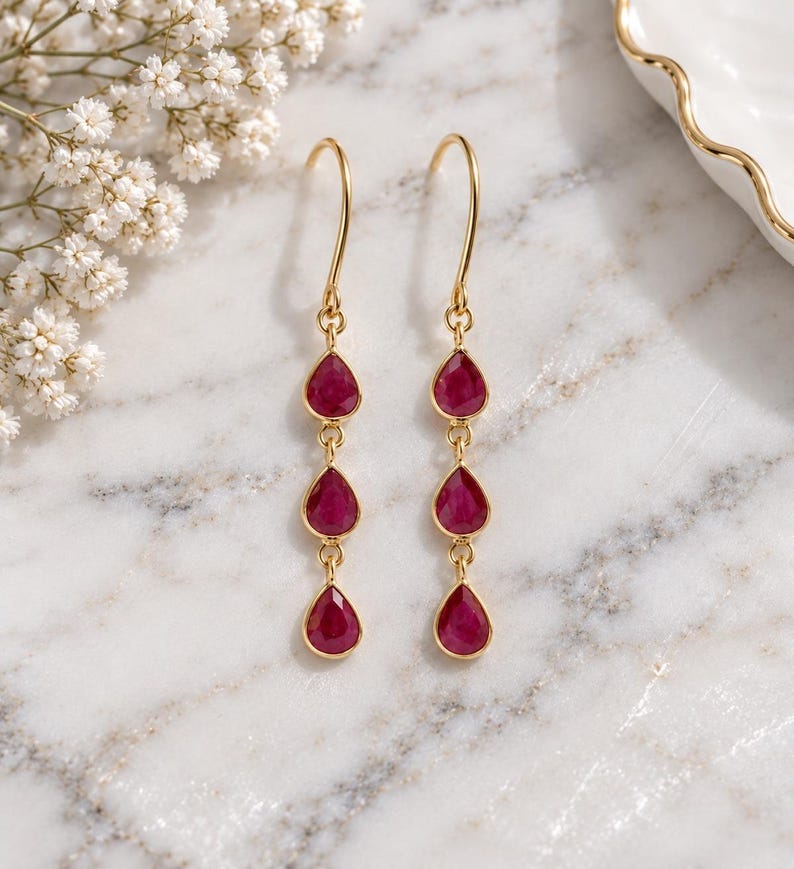 May include: A pair of gold drop earrings, each featuring three vertically aligned, teardrop-shaped, deep pinkish-red gemstones suspended from a curved gold ear wire. The stones are set in thin gold bezels and connected by small gold jump rings.