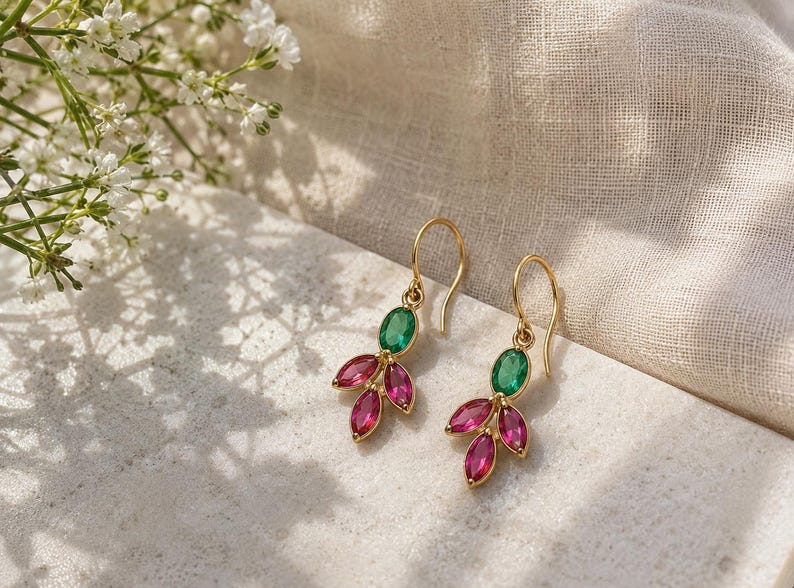 May include: A pair of gold drop earrings featuring a single oval-cut green gemstone at the top, accented by three marquise-cut pink gemstones arranged in a leaf-like cluster at the bottom, suspended from simple gold ear wires.