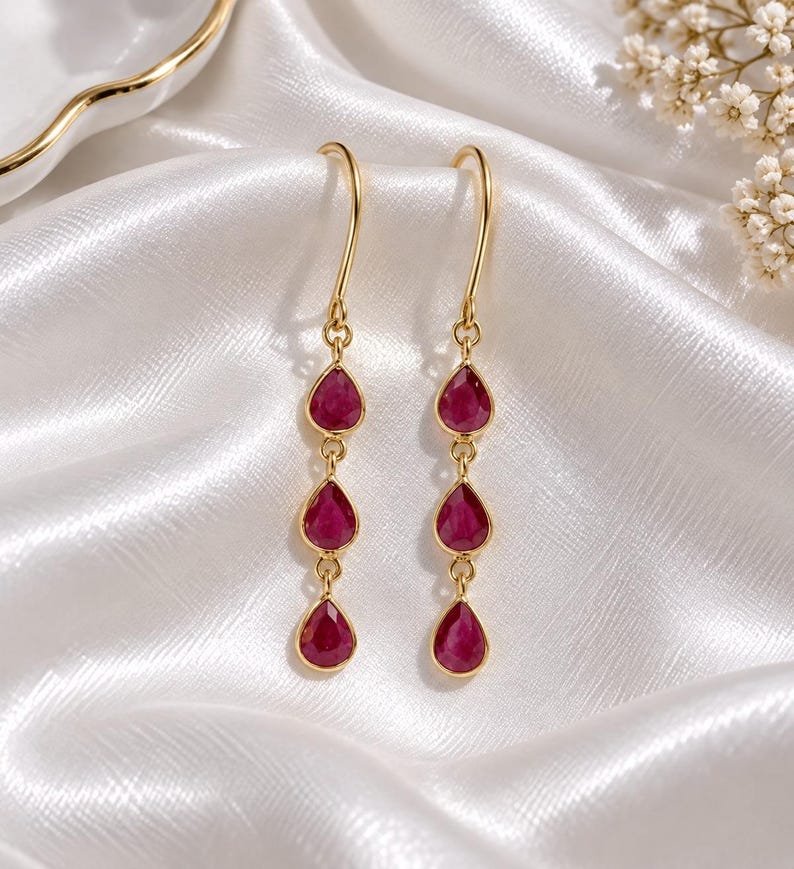 May include: A pair of gold drop earrings featuring three vertically aligned, teardrop-shaped, deep pink gemstones suspended from a curved gold hook. The earrings have a polished metallic finish and a delicate, articulated design that allows for movement.