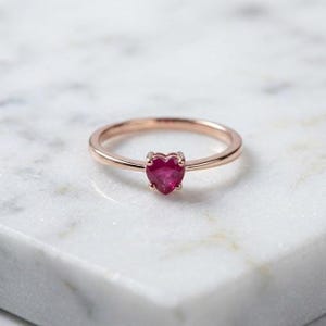 Può includere: Un anello in oro rosa con un rubino a forma di cuore. L'anello ha una fascia sottile e un design semplice. Il rubino è di un rosso vibrante, incastonato in una montatura a quattro griffe. L'anello è esposto su una superficie di marmo bianco.