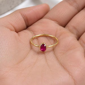 May include: A thin gold band ring featuring a single, small, oval-shaped, faceted ruby gemstone set in a bezel setting. The ring has a polished, smooth finish and a minimalist design, showcasing the vibrant, deep red color of the central stone.
