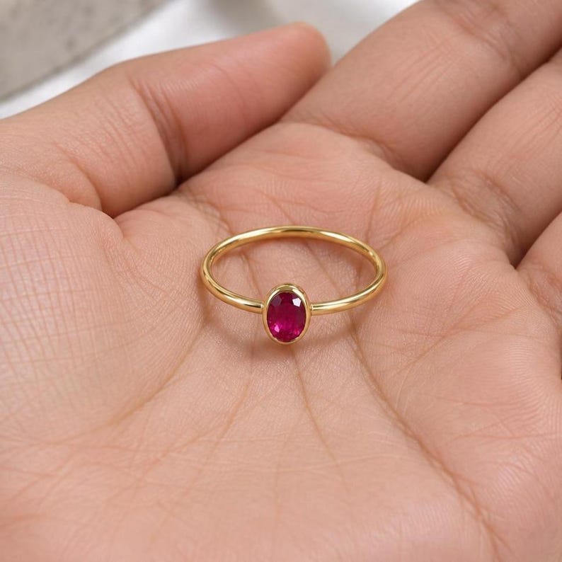 May include: A thin gold band ring featuring a single, small, oval-shaped, faceted ruby gemstone set in a bezel setting. The ring has a polished, smooth finish and a minimalist design, showcasing the vibrant, deep red color of the central stone.