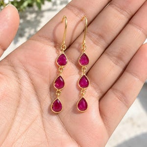 May include: A pair of gold drop earrings, each featuring three vertically aligned, teardrop-shaped, vibrant magenta-pink gemstones. The earrings are suspended from simple gold wire hooks, creating a delicate and elongated design with a polished finish.