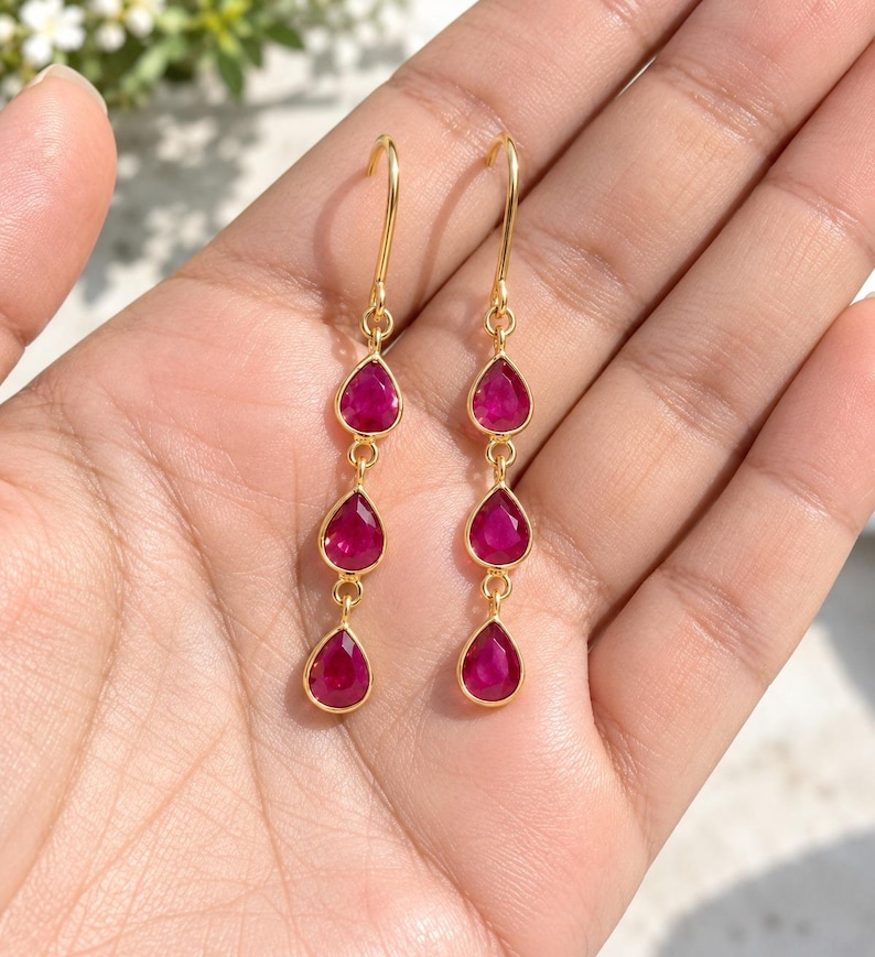 May include: A pair of gold drop earrings, each featuring three vertically aligned, teardrop-shaped, vibrant magenta-pink gemstones. The earrings are suspended from simple gold wire hooks, creating a delicate and elongated design with a polished finish.