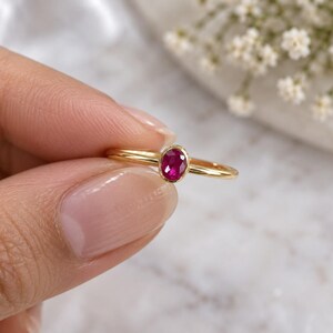 May include: A gold ring featuring a small, oval-cut, vibrant pinkish-red ruby gemstone set in a simple bezel setting. The band is thin, polished, and circular, showcasing a minimalist design that highlights the central gemstone's deep color.