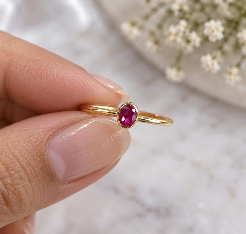 May include: A gold ring featuring a small, oval-cut, vibrant pinkish-red ruby gemstone set in a simple bezel setting. The band is thin, polished, and circular, showcasing a minimalist design that highlights the central gemstone's deep color.
