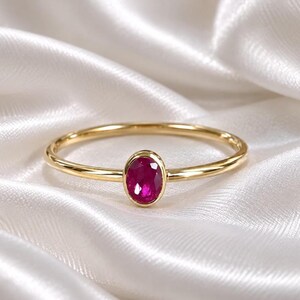 May include: A thin gold band ring featuring a single oval-shaped, faceted deep pink ruby gemstone set in a smooth bezel setting. The ring has a polished metallic finish and a simple, slender design that highlights the vibrant color of the stone.