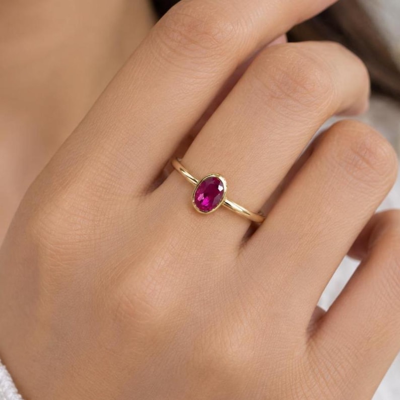 May include: A gold ring featuring a single oval-shaped, deep pinkish-red gemstone set in a smooth bezel setting. The band is thin and polished, showcasing a simple design that highlights the vibrant color and clarity of the central stone.