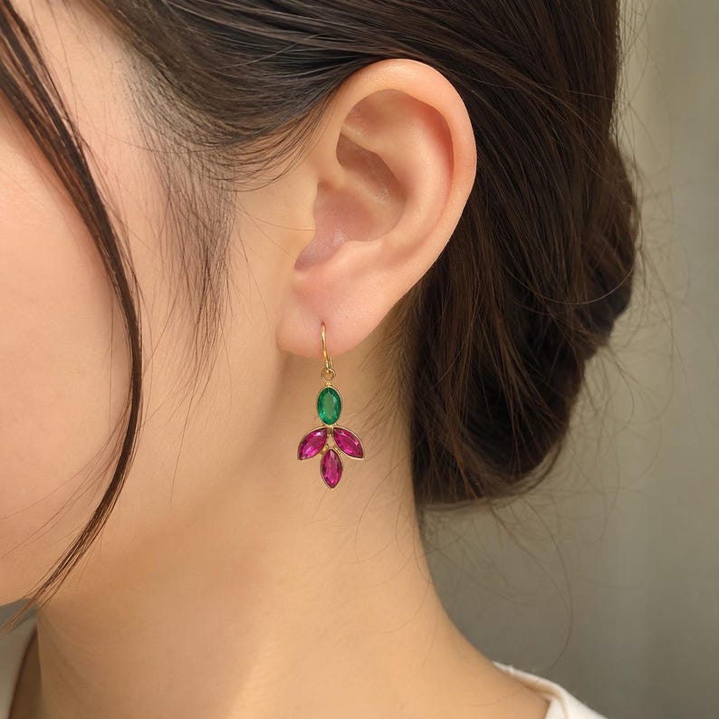 May include: A gold-toned drop earring featuring a single oval-shaped green gemstone at the top, suspended above three marquise-cut fuchsia pink gemstones arranged in a fan-like cluster. The earring hangs from a simple gold-toned hook wire.