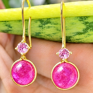 Peut inclure: Une paire de boucles d'oreilles pendantes dorées avec une pierre ronde facettée rose vif suspendue à une pierre plus petite, ronde et facettée rose clair. Les boucles sont fixées à des crochets dorés pour oreilles percées.