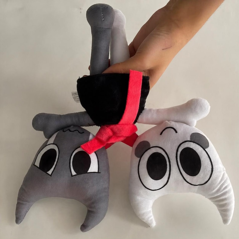 Op de afbeelding: Een knuffel met een zwarte kop, een rode sjaal en grijze en witte ledematen. Het speelgoed heeft drie cartoonachtige gezichten, &eacute;&eacute;n grijs en twee wit, elk met grote ogen. Het speelgoed wordt door een hand tegen een witte achtergrond gehouden.
