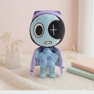 May include: A plush toy featuring a light blue, round head with one large white eye and one black eye patch with a white cross. It wears a purple cape and holds a light blue, star-shaped object with purple spots. The character has a small mouth.