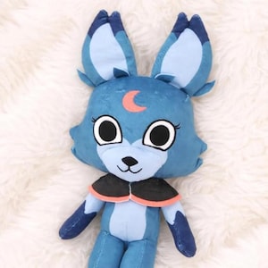 May include: A plush toy fox with large ears and a crescent moon design. The fox is primarily blue with black and orange accents. It has large, round eyes and a small black nose. The toy is made of soft material and is designed to be cuddly.