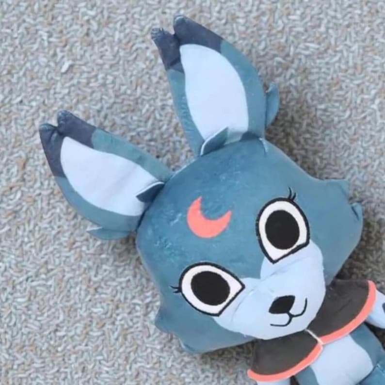 May include: A plush toy in the shape of a blue fox with large ears and a crescent moon detail. The fox has large, round eyes, a black nose, and a black and orange collar. The toy is made of soft fabric and is designed for children.