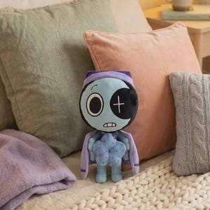 May include: A plush toy featuring a light blue, round head with a large white eye on the left and a black eye patch with a white cross on the right. It has a small mouth, a purple hood, and a light blue body holding a star-shaped object.