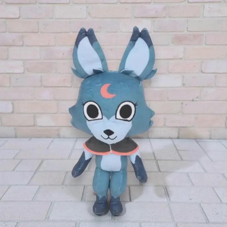 May include: A plush toy in the shape of a blue fox-like creature with large ears. It has a crescent moon design on its forehead and a black and orange collar. The toy is standing on a brick surface.