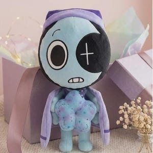 May include: A plush toy featuring a light blue, round head with one large white eye and one black eye patch marked with a white plus sign. The figure has a light blue body, a purple cape, and holds a textured, light blue star-shaped object in its hands.