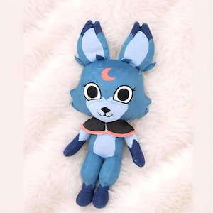 May include: A plush toy fox in shades of blue, with large ears and a crescent moon detail on its forehead. The fox has large, round eyes, a black nose, and a black and orange collar. The toy is made of soft fabric and is designed to be cuddly.