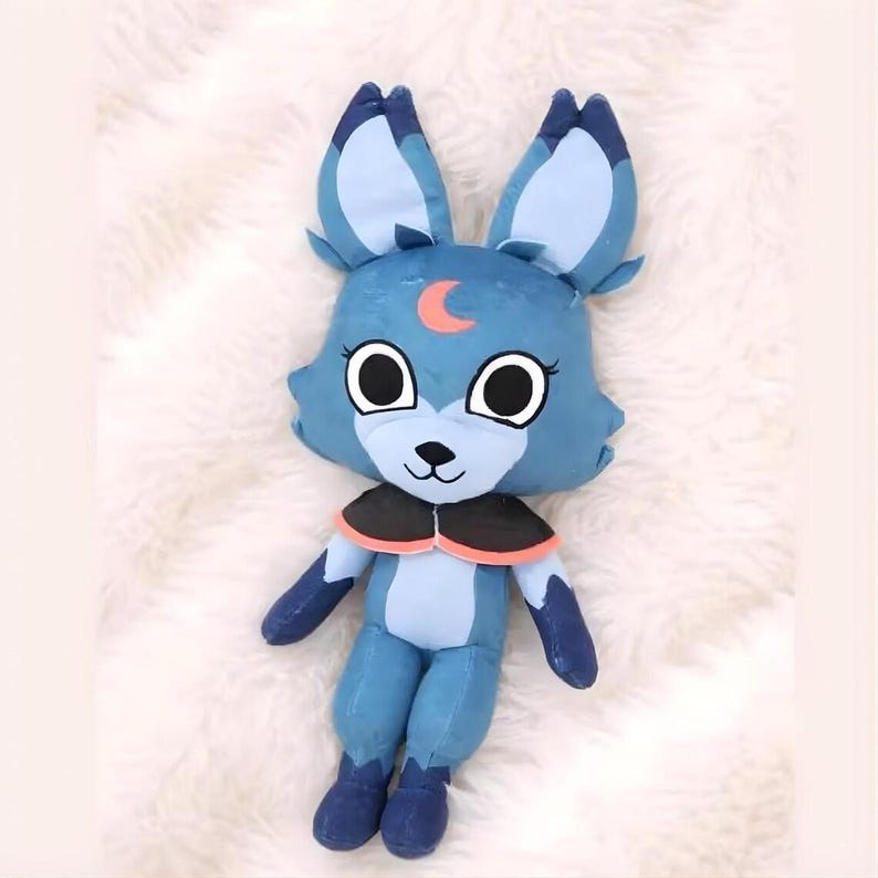 May include: A plush toy fox in shades of blue, with large ears and a crescent moon detail on its forehead. The fox has large, round eyes, a black nose, and a black and orange collar. The toy is made of soft fabric and is designed to be cuddly.