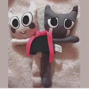 Op de afbeelding: Twee pluche kattenpoppen. E&eacute;n is wit met een lachend gezicht en de andere is grijs met een verdrietig gezicht. Ze zijn verbonden door een roze sjaal. De poppen hebben grote, ronde ogen en zijn gemaakt van zachte stof.