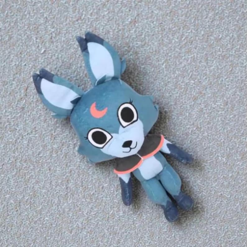 May include: A plush toy fox with large eyes, a crescent moon detail, and a small, dark brown collar. The fox is primarily blue with white and orange accents. The toy is lying on a textured, light gray surface.