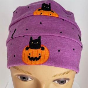 May include: Purple beanie with a black cat inside a jack-o'-lantern pattern. The beanie has a fold-over brim.