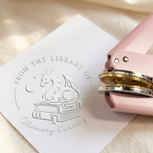 Peut inclure: Papier blanc gaufré avec un motif de dragon, le texte "FROM THE LIBRARY OF Memory Carving" et une pince à gaufrer rose. Le dragon est perché sur une pile de livres, avec une lune et une étoile.