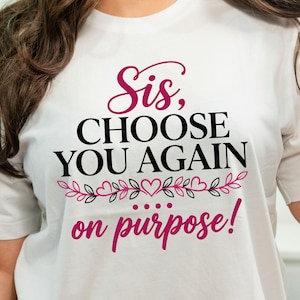 Può includere: T-shirt bianco con la scritta "Sis, CHOOSE YOU AGAIN on purpose!" in nero e rosa. Il design include una grafica floreale e a forma di cuore. Un capo d'abbigliamento casual.