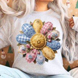 May include: A light gray sweatshirt featuring a large floral design. The flower has petals in various patterns, including gingham, lace, and floral prints, in shades of blue, pink, and yellow. The center of the flower is gold, with a button detail.