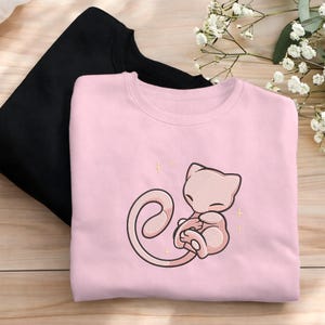 Könnte beinhalten: Zwei Sweatshirts, eines in Schwarz und eines in Hellrosa. Das rosa Sweatshirt hat ein gesticktes Design eines hellrosa Cartoon-Charakters. Der Charakter ist zusammengerollt. Das Design ist mit kleinen goldenen Sternen akzentuiert.