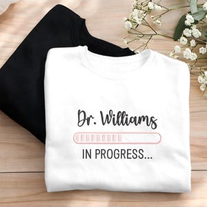 May include: Two sweatshirts, one black and one white, are displayed. The white sweatshirt features the text "Dr. Williams" above a loading bar graphic and the words "IN PROGRESS..." in black font. The black sweatshirt is folded behind the white one.