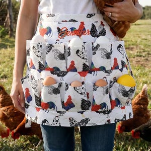 May include: A white apron with multiple pockets, decorated with rooster and hen illustrations in red, black, and blue. The apron is filled with eggs of various colors. A brown hen is held in the arms. The word "Jennifer" is embroidered on the apron.