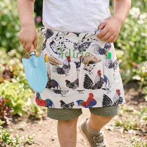 May include: A white waist apron featuring a repeating pattern of colorful roosters and chickens, with multiple pockets containing twine and tools. The name Felicia is embroidered in green script on the front center of the apron fabric.