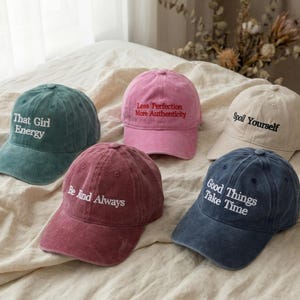 Könnte beinhalten: Fünf Baseballkappen in verschiedenen Farben: Türkis, Rosa, Beige, Burgunderrot und Marineblau. Jede Kappe hat einen gestickten Text: "That Girl Energy", "Less Perfection More Authenticity", "Spoil Yourself", "Be Kind Always" und "Good Things Take Time."