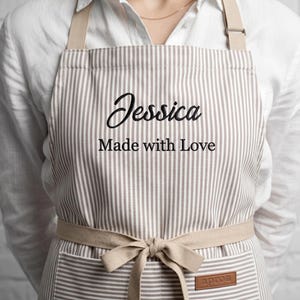 Könnte beinhalten: Eine beigeweiß gestreifte Schürze mit dem eingestickten Namen Jessica und dem Text Made with Love in schwarzer Schrift. Die Schürze verfügt über beigefarbene Stoffbänder an der Taille, die zu einer Schleife gebunden sind, und ein kleines braunes Etikett.