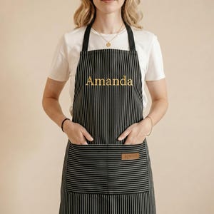 Op de afbeelding: Zwart-wit gestreepte schort met de naam "Amanda" in goud geborduurd. De schort heeft een klein bruin label met het woord "apron". Het heeft zakken en een nekband.