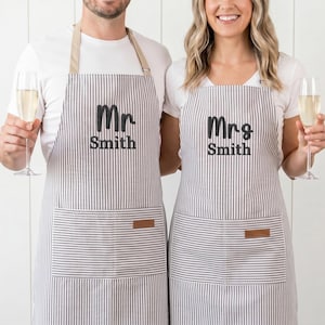 以下が含まれることがあります： ストライプ柄のお揃いのエプロン。片方には「Mr Smith」、もう片方には黒で「Mrs Smith」とプリントされています。エプロンにはポケットと革のパッチが付いています。カップルはシャンパングラスを持っています。