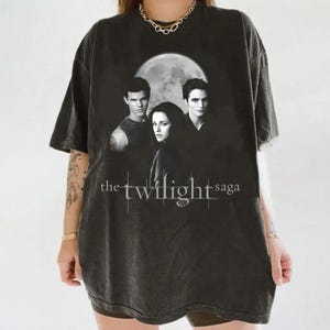 May include: Dark gray t-shirt featuring a graphic of three people in front of a full moon, with the text "the twilight saga" in white. The shirt has a vintage, worn look.