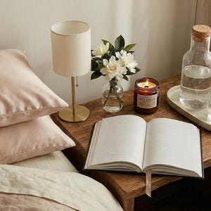 May include: Two light pink satin pillowcases on a bed next to a wooden nightstand holding a small lamp, a glass vase with white flowers, a lit candle, an open notebook, and a glass carafe with a glass of water on a ceramic tray.