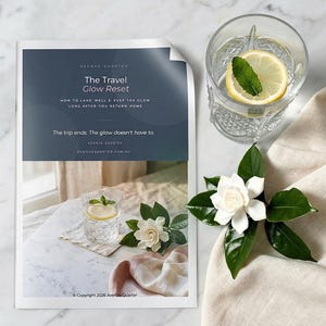 May include: A printed guide titled The Travel Glow Reset with text about landing well and keeping the glow. Beside it sits a clear glass of water with a lemon slice and mint leaf, and a white gardenia flower on a beige fabric cloth.