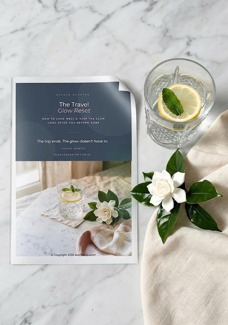 May include: A printed guide titled The Travel Glow Reset with text about landing well and keeping the glow. Beside it sits a clear glass of water with a lemon slice and mint leaf, and a white gardenia flower on a beige fabric cloth.