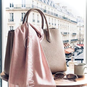 May include: A dusty pink scarf with the white cursive text Avenue draped over a structured beige leather handbag. Beside the bag are brown sunglasses, a small glass perfume bottle, and a ceramic mug on a round wooden table surface.