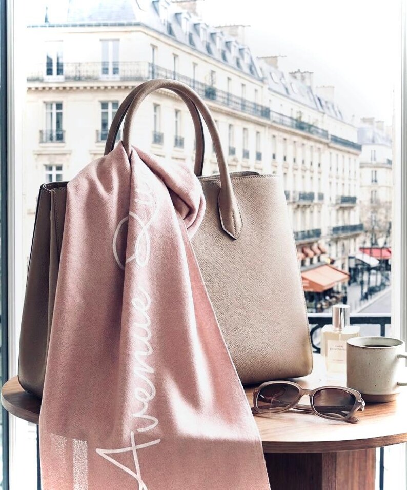 May include: A dusty pink scarf with the white cursive text Avenue draped over a structured beige leather handbag. Beside the bag are brown sunglasses, a small glass perfume bottle, and a ceramic mug on a round wooden table surface.