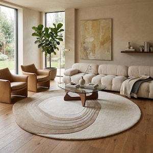May include: A large, round area rug featuring a concentric, swirling pattern in shades of cream, beige, and light brown. The rug is placed on a wooden floor in a room with two tan leather armchairs, a cream-coloured sofa, and a glass coffee table.